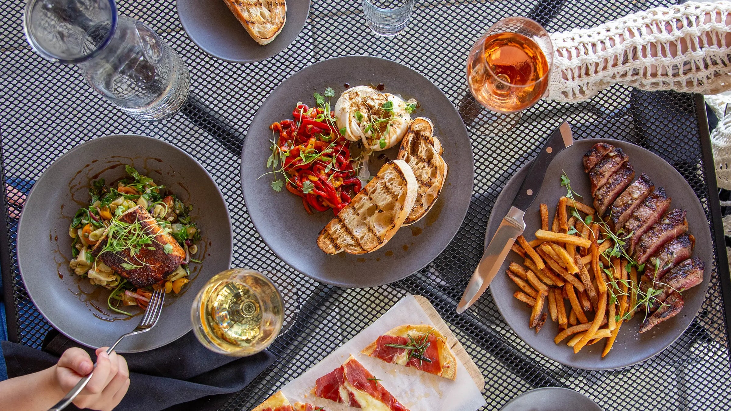 Plated dishes of buratta, steak frites, flatbread and two glasses of wine on a patio table.