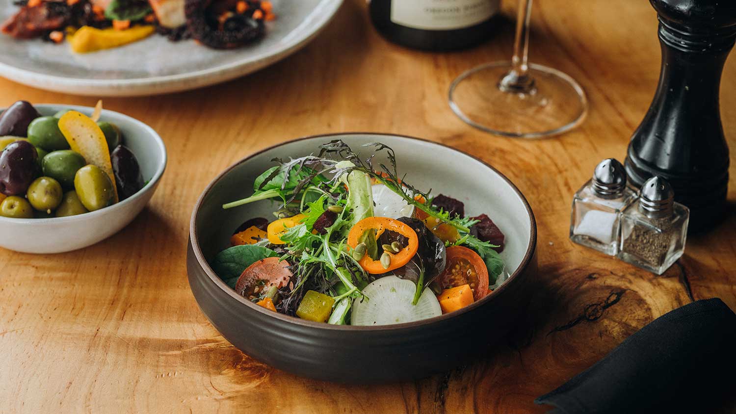 Salad with spring mix, orange bell pepper, tomatoes, and radishes with a bowl of olives in the background