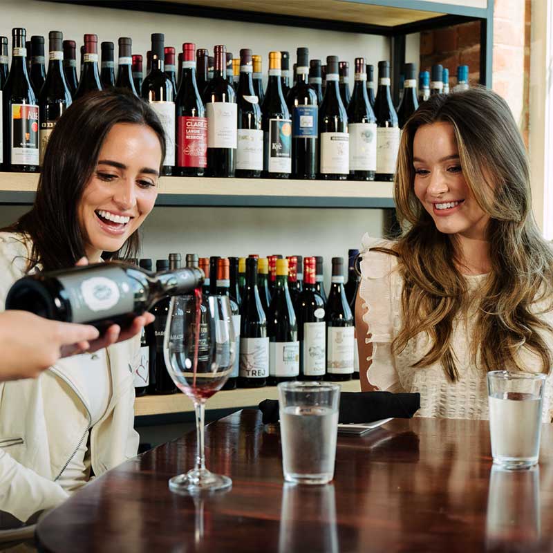 Server pours two women red wine at a table by the window.