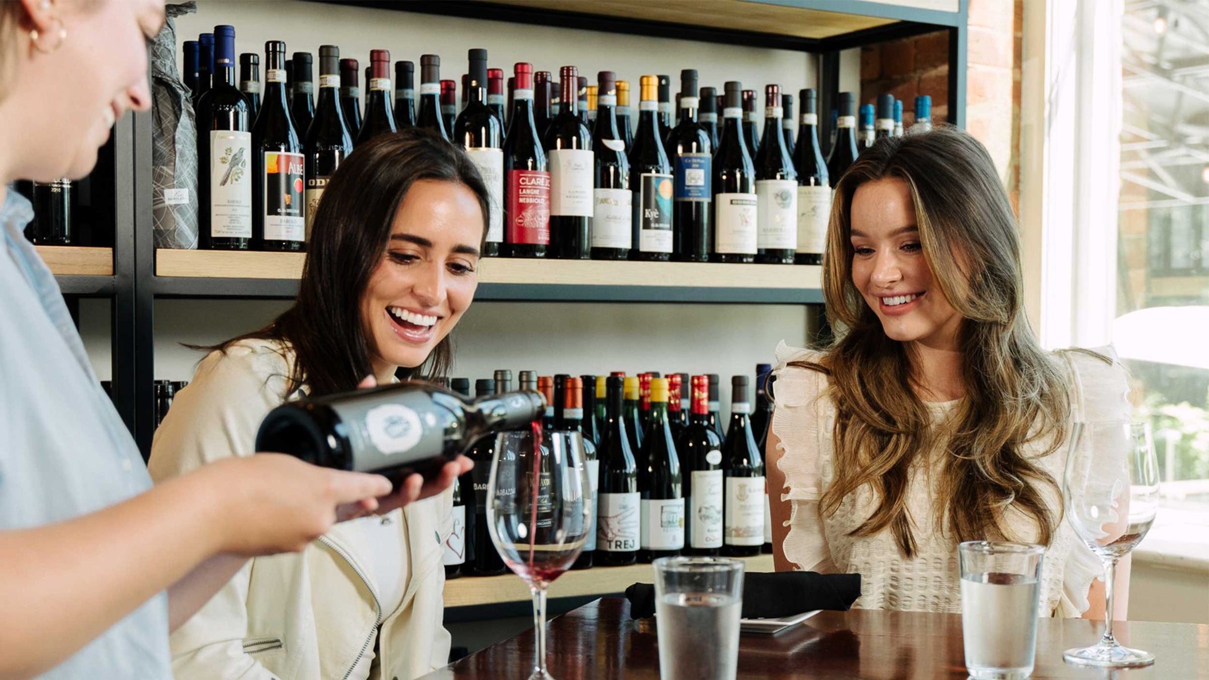 Server pours two women red wine at a table by the window. 